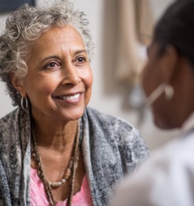 An older woman with gray curly hair smiles while talking to another person in a well-lit indoor setting.