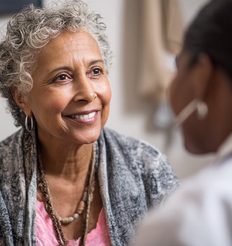 An older woman with gray curly hair smiles while talking to another person in a well-lit indoor setting.