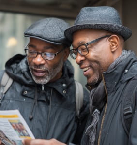 Two men wearing glasses, hats, and jackets stand close together outdoors, looking at and discussing a brochure or pamphlet.