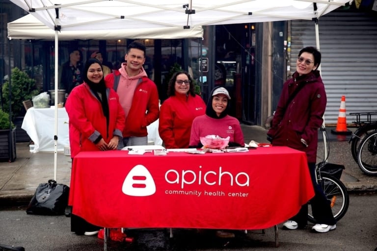 Five people stand behind and beside a table with an "apicha community health center" banner at an outdoor event, under a white canopy, with informational materials on the table.