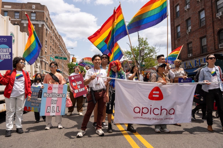 A group of people hold rainbow flags and signs supporting healthcare and LGBTQ+ rights at a parade; a large banner reads "apicha community health center.