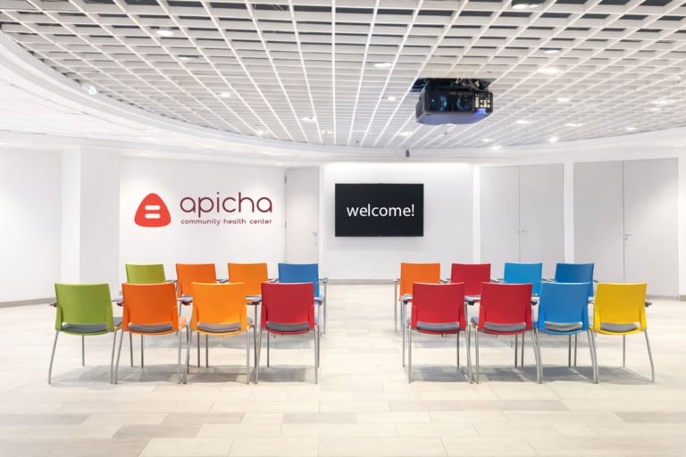 A conference room with colorful chairs facing a screen that displays "welcome!" and a wall with the Apicha Community Health Center logo.