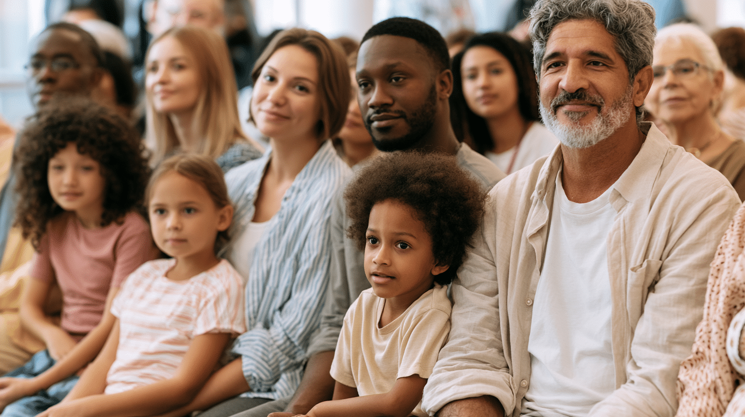 A diverse group of adults and children sit closely together, attentively watching something in front of them in a bright indoor setting.