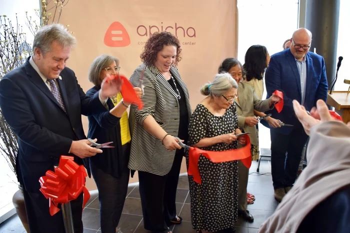 A group of people stand together cutting a red ribbon at an indoor event in front of a backdrop that reads "apicha community health center.