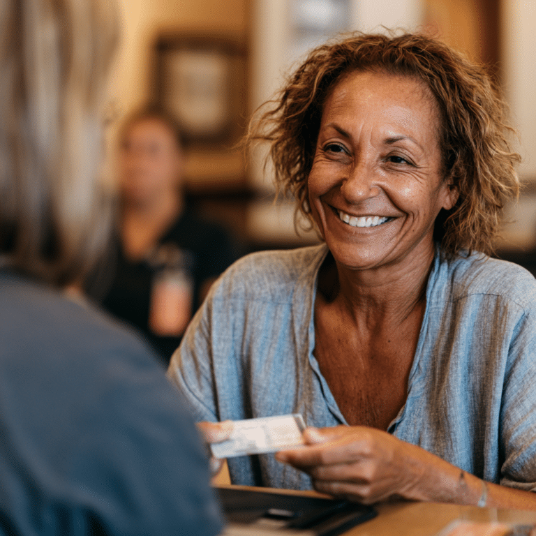 A woman smiles while handing over an identification card to another person across a table in an indoor setting.