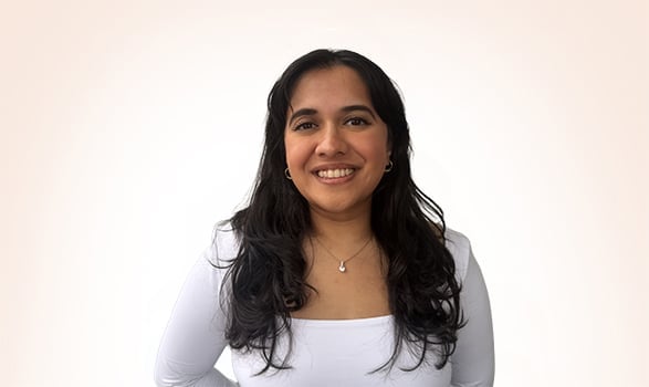 A woman with long dark hair, wearing a white top and necklace, stands in front of a plain light background, smiling at the camera.