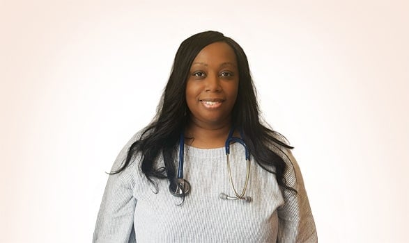 Woman wearing a light gray top and a blue stethoscope around her neck, standing against a plain light background, smiling at the camera.