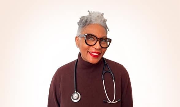 Smiling person with gray hair wearing glasses, a maroon turtleneck, and a stethoscope around their neck stands against a plain, light background.