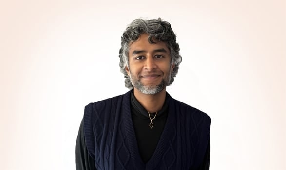 A person with short, curly gray hair and a beard, wearing a dark sweater vest and a gold necklace, stands facing the camera against a plain light background.